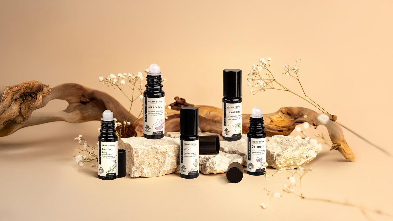 Aroma Tierra essential oil roll-ons — Breathe Easy, Sleep Aid, Joy, Head Lite and De-Stress — arranged on natural stone and driftwood with dried flowers